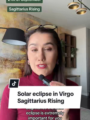 A woman with dark hair and a red turtleneck is holding a microphone and talking. The text overlay reads, "Solar eclipse in Virgo Sagittarius Rising". The background consists of a living room setting.