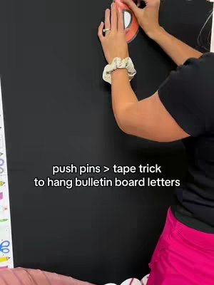 A person's hands are shown placing a letter template onto black paper affixed to a wall. The person, wearing a scrunchie and a ring, holds the template, which is white with an adjacent orange border. The text overlay reads: 'push pins > tape trick to hang bulletin board letters'. In the background, a white and green section of a bulletin board is visible.