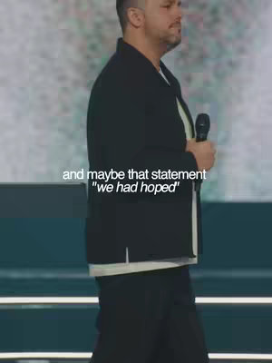 A man in a black jacket and white shirt stands on a stage with a microphone in hand. Text overlay appears on the image.