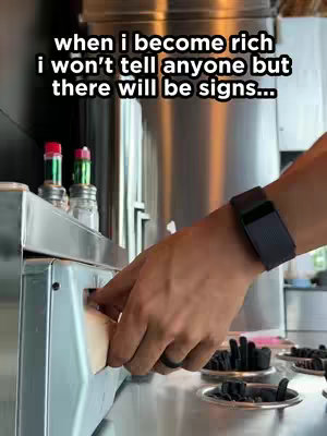 A hand wearing a black watch and a ring is reaching into a napkin dispenser in a restaurant setting. The background shows stainless steel equipment and other restaurant supplies. Text overlayed on the image says 'when i become rich i won't tell anyone but there will be signs...'.