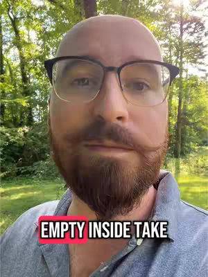 A close-up shot of a man's face. He is wearing glasses and has a bald head, with a full beard and mustache. The background is a blur of green trees. The foreground includes a text overlay that says 'EMPTY INSIDE TAKE'.