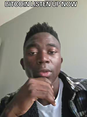 A selfie-style video cover image featuring a Black man with short, textured hair and a neutral facial expression. He has a slight smirk and is resting his chin on his left hand. He wears a flannel shirt over a white shirt and looks directly at the camera against a blurred background. The text overlay across the top of the image reads "BITCOIN LISTEN UP NOW".
