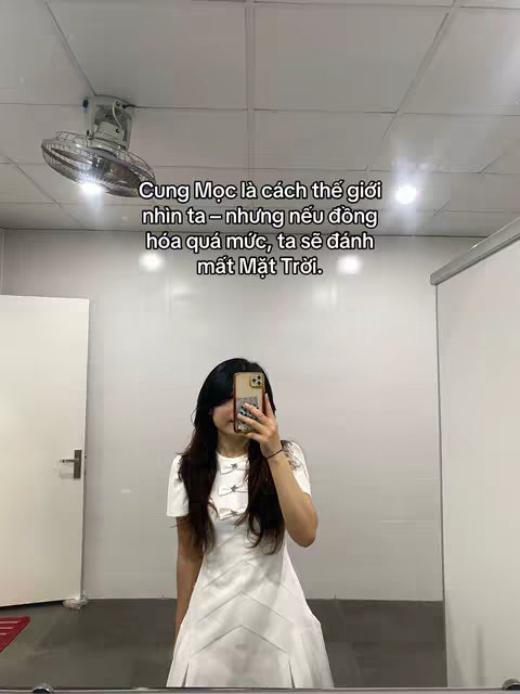 A person is taking a selfie in front of a mirror, showing the upper body and the environment. They are in a room that appears to be a bathroom or a utility space, indicated by the white walls and a ceiling fan. The person is wearing a white dress. Text in Vietnamese is overlaid on the image.