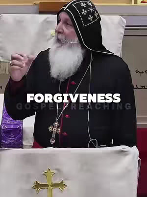A medium shot features a man in religious garb speaking at a podium. The man has a white beard, and is wearing a black robe over a red under-garment. A black hood and cap complete the attire. Text overlays partially cover him. The background has a white wall, with some text to the right, and a gold cross on the bottom right of the image, with a purple background.