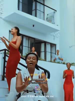 The image showcases a group of four women standing and singing in front of a white building with a balcony. The woman in the center is sitting in a chair, singing with her hands gesturing, and there is text overlayed on the image for the video's title and other information.