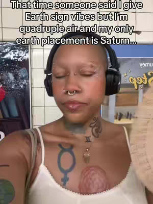 The cover image features a close-up of a person with a shaved head, multiple facial piercings, and headphones around their neck. Text is overlaid at the top of the image that reads, "That time someone said I give Earth sign vibes but I'm quadruple air and my only earth placement is Saturn..." The person has a tattoo on their chest and is making eye contact with the viewer, indicating a personal and conversational style.
