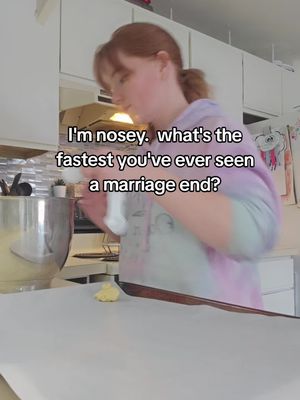 The image shows a woman in a kitchen preparing what seems to be cookie dough. The woman is wearing a light-colored tie-dye sweatshirt with her red hair pulled back in a ponytail. She is focused on forming dough on a sheet of parchment paper. The background includes kitchen cabinets and cookware.