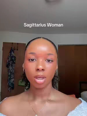 A close-up shot of a woman with dark skin. She has dark, neatly styled hair and is wearing makeup. The background is out of focus showing a room. Text overlay reads "Sagittarius Woman".