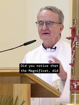 The image shows a man speaking into a microphone at a lectern. He is wearing a white clerical collar and glasses. The background is a plain, off-white wall. There is a banner to the right with the letters "A" and "5" on it. Overlay text at the bottom of the screen reads: "Did you notice that the Magnificat, did".