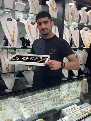A man stands behind a jewelry display case holding a tray with the word 'SORRY' spelled out in gold-colored jewelry. The background shows numerous necklaces and jewelry displays.