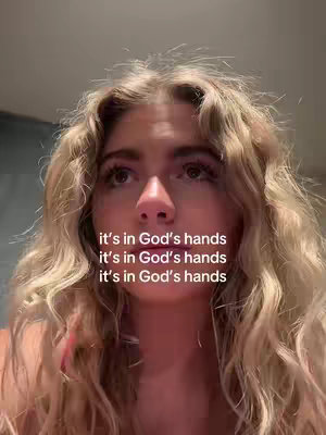 A close-up shot of a young woman with long, wavy blonde hair looking upwards with a contemplative expression. Text overlays repeat the phrase "it's in God's hands". The background is out of focus.