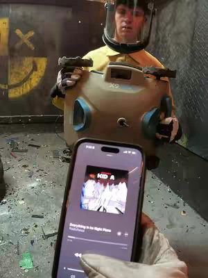 A first-person perspective shot, someone is holding a phone showing an album cover and is also holding a device while wearing protective gear, and is standing in a room with broken debris on the floor.