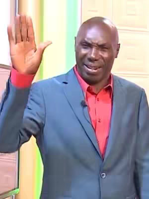 A man wearing a red shirt and a blue blazer, raising his right hand with his palm facing forward. His facial expression appears concerned or questioning. The background is a mix of green and white.