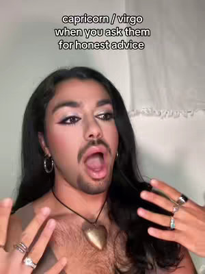 The image is a selfie-style shot of a person with full makeup and accessories, a heart pendant, and multiple rings. They are looking at the camera with an open mouth, with the text overlayed on the top. The background is neutral. The color palette is natural skin tones combined with bold makeup.