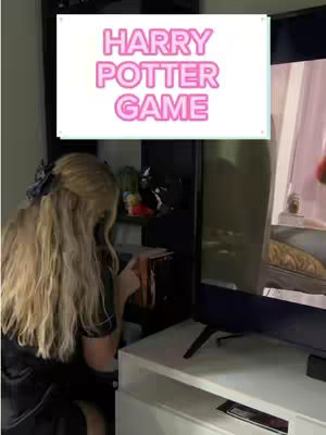 A person is sitting, facing away from the camera, with long blonde hair. The person is leaning towards a TV screen on the right. A sign with the text "HARRY POTTER GAME" in pink letters is overlaid in front of the screen in the background.