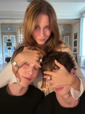 A woman stands behind two people and covers their eyes with her hands. The background shows a room.