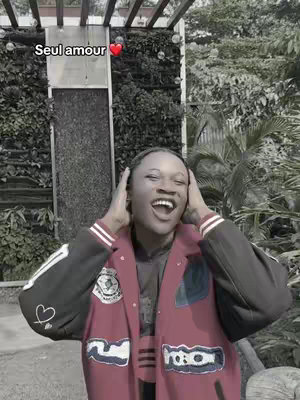 A woman with a surprised expression holds her hands to her head. She wears a burgundy jacket and stands in front of a background of greenery.