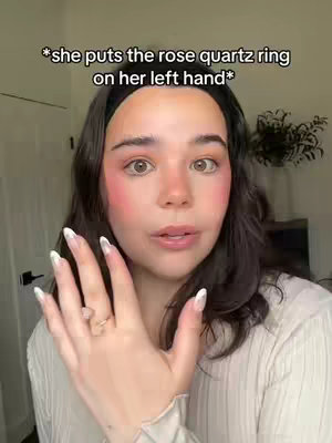 A woman with dark hair and subtle makeup looks directly at the camera, gesturing with her left hand. A ring with a small, light-colored stone is visible on her ring finger. She wears a white, ribbed, long-sleeved shirt and a black headband. The background is softly lit, suggesting an indoor setting.