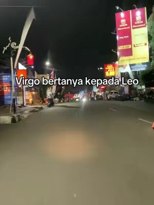 A nighttime street scene, likely filmed from inside a vehicle, with a blurred road leading towards distant lights. The sky is dark with street lights and building lights illuminating the area. Text overlayed in the image reads "Virgo bertanya kepada Leo".