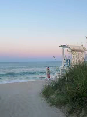 A serene beach scene at dusk with a pathway leading towards a lifeguard station. The sky transitions from a soft blue to a pale pink, reflecting on the water's surface. The beach is golden, with green sea grass and a white lifeguard station. The perspective is low, creating a sense of depth.