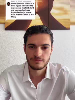 A man is centered in the frame, looking directly at the camera with a neutral expression. He has short, dark hair and a trimmed beard. He is wearing a white button-down shirt against a plain white wall background. Above him, a comment box is visible at the top with a comment from 'Jetmir' set against a blurred backdrop featuring a cross within a golden light, suggesting religious imagery.