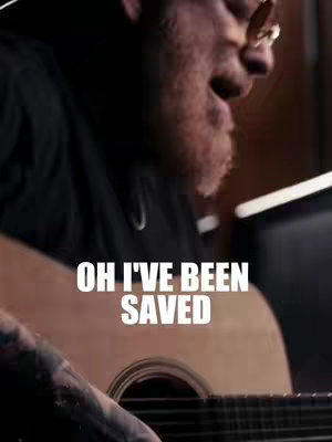The image captures a man with a beard and glasses playing an acoustic guitar. The shot is from a slightly low angle, focusing on the man's face, guitar, and the text overlayed. The lighting appears to be natural, and the overall feel is warm and inviting.