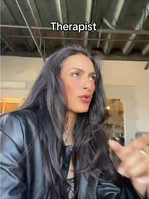 A selfie-style shot of a woman speaking. She has long black hair and is wearing a black leather jacket. The background includes architectural details like exposed beams and what appears to be a wooden wall, suggesting an indoor setting with a vintage or industrial design.