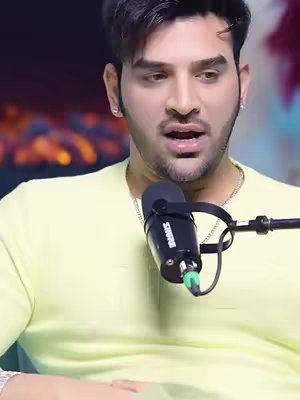A man with dark hair and a light yellow shirt is centered in this shot, with a microphone close to his mouth. The background is slightly out of focus. The man has light skin and is looking down and slightly to the side with an open mouth. A microphone is positioned in front of him.
