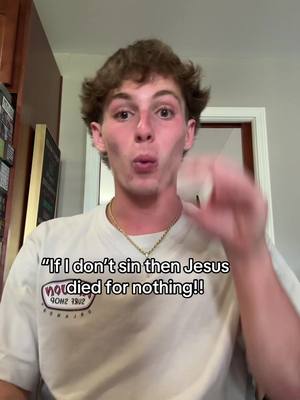 A young man with short, wavy brown hair is the central focus, positioned against a home interior backdrop. He's wearing a white t-shirt and a gold chain necklace, with text overlaying the shirt. The background includes a plain gray wall to the left and a slightly blurred doorway on the right, suggesting a domestic setting. The lighting appears natural, and the overall composition is a medium shot, emphasizing the man's face and upper body. His pose is animated; he gestures with his right hand as if making a point and has a slightly puckered mouth.