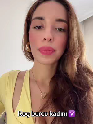 A woman with light skin and long brown hair appears in a selfie-style shot. She wears a yellow top and gold hoop earrings and has rosy cheeks and pink lipstick. The text overlay at the bottom reads, "Koç burcu kadını" with aries symbol next to it