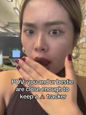 A close-up shot featuring a woman with fair skin, dark hair, and bold makeup. She is covering her mouth with her hand, eyes wide as if in surprise. The background is a blurred interior space. A text overlay at the bottom reads, "POV: you and ur bestie are close enough to keep a 💩 tracker".
