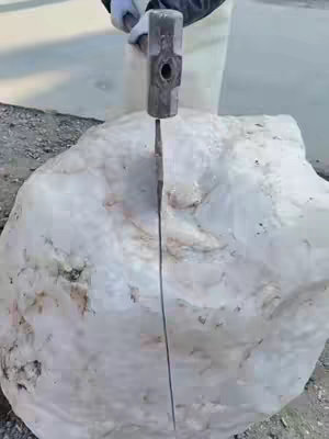 A large, uncut white crystal is being split using a large hammer and chisel. The crystal is in the center of the image, positioned against a background of what appears to be concrete.
