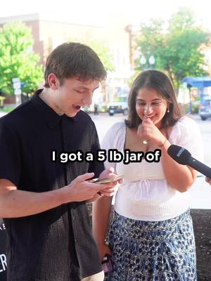 A man and a woman are standing outside, with the man holding a phone and looking at it with a surprised expression, the woman is smiling with the microphone pointed at them.