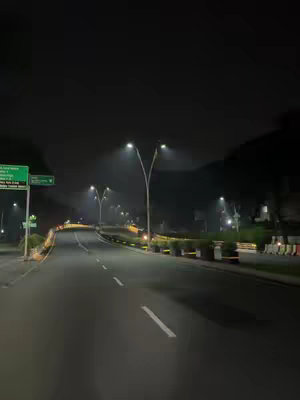 A nighttime shot of a road curving into the distance, lit by streetlights. The road is dark, with visible lane markings, leading towards a darker sky. Streetlights line the sides of the road.
