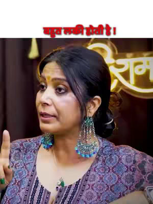 A woman with dark hair, wearing a patterned top and large earrings, is pointing upwards and speaking. Text in Hindi is overlaid at the top of the image.