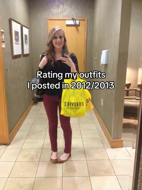 A woman stands in a well-lit room wearing a black top, burgundy pants, and flats. She holds a phone in one hand and a yellow Forever 21 shopping bag. The room is decorated with framed pictures and a mirror.