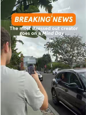 A person is holding a phone in front of them while standing in front of a street scene The phone is pointed towards a black car in the street, in the background there is a street with buildings and trees in the background.