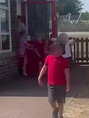 A young child, blurred out, walks toward the camera with a red shirt and dark shorts. Other children blurrily exit the school building behind them through a red door and the building structure is also blurred. The environment looks like a school, and a metallic fence is visible on the right side of the frame.