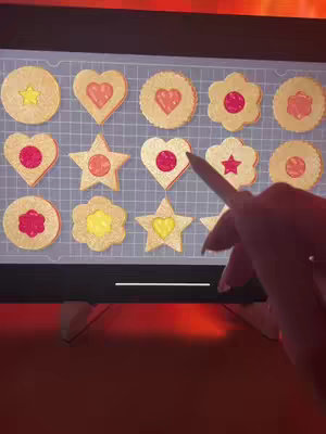 An iPad screen displays a grid of various cookie-shaped puzzles. A hand holding a stylus is pointing towards the puzzle elements. The lighting is warm, and the composition has a close-up perspective.