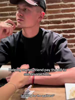 A man wearing a black shirt and a baseball cap is seated at a table, looking towards the left. Another person's hand with a bracelet is partially visible in the foreground, as well as a section of the table. Text overlays appear on the image.