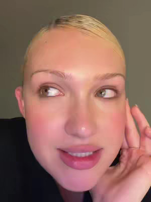 A close-up shot of a woman's face, slightly angled, with a light-skinned complexion and blonde hair. Her eyes are looking to the side, and she is smiling slightly while holding her hand near her face. The background is a muted grey-green color, emphasizing the subject's features.