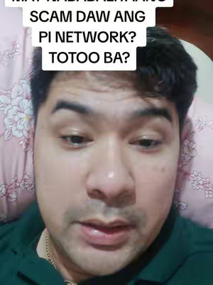 A close-up shot of a man in a green shirt, lying on a bed with a floral-patterned pillow. Above his head, a text overlay poses a question regarding the legitimacy of Pi Network. The man has dark hair and is looking down slightly as if he's speaking or thinking, with a neutral expression.
