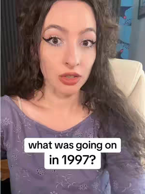 A woman with curly dark hair is in a close-up shot, looking directly at the camera with an expressive face. She wears a pale purple top with subtle floral embroidery around the neckline. A white text box at the bottom of the frame reads, "what was going on in 1997?"