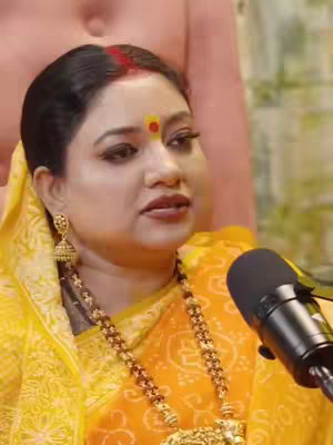 A woman in a yellow saree is centered in the frame, speaking near a microphone. She has dark hair pulled back, a red mark on her forehead, and a yellow dot in the middle. Gold earrings and a long gold necklace adorn her. A microphone is positioned to her right.