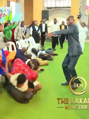 A man in a suit gestures towards a group of people lying on the floor in a large room with other people standing or sitting nearby. In the foreground, there are people lying on the bright green floor, and in the background are people on chairs. In the bottom right corner is a golden logo with the text "TKM" with the words "THE KAKANDE MINISTRIES" below it.