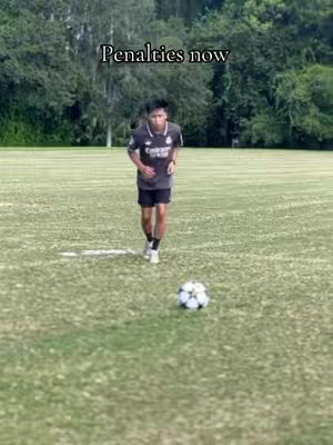 A young man in a dark jersey is running towards a soccer ball on a green field, the text overlay 'Penalties now' is clearly visible at the top of the image.