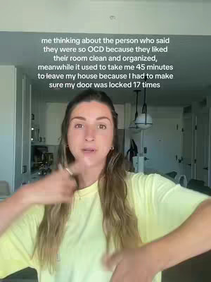 A woman with long brown hair is in a well-lit interior space, likely a home. The background features a kitchen area with white cabinets and a glimpse of hanging light fixtures. The woman is wearing a light yellow short-sleeved shirt. She gestures with her hands as she speaks, with a serious expression on her face.
