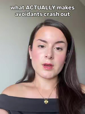 The image is a selfie-style shot featuring a woman from the chest up. She has dark hair, fair skin, and light makeup including blush and lip color. She's wearing a black off-the-shoulder top and a gold necklace with a heart pendant. The background appears to be a plain wall. The text overlay in the image states: "what ACTUALLY makes avoidants crash out".