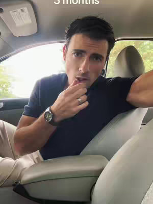 A man is seated in the driver's seat of a car, looking directly at the camera with a thoughtful expression. He's wearing a black shirt, a watch, and a ring. The interior of the car is visible, with the seat, headrest, and part of the windshield in the background.