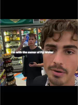 A selfie-style video captured in a convenience store. The main subject, a man with a mustache and curly hair, takes up the right side of the frame. Behind him, another man holds up a Fiji Water bottle. The background shows store shelves with various products and a beverage cooler.